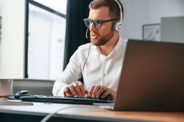 In headphones. Man in formal clothes is working in the modern office. Using computer