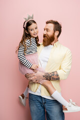 Tattooed man holding daughter with crown headband on pink background.