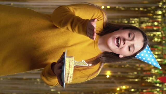 Beautiful happy young woman standing over golden background, holding a birthday cake with number 1 candle, looking at camera at home party. POV. Birthday holiday party people emotions.