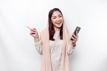 Excited Asian woman wearing hijab pointing at the copy space beside her while holding her phone, isolated by white background