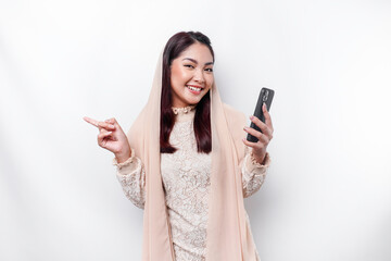 Fototapeta premium Excited Asian woman wearing hijab pointing at the copy space beside her while holding her phone, isolated by white background