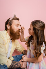 Preteen girl applying eyeshadow on tattooed father with crown headband on pink background.