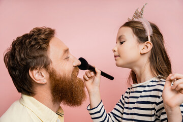 Side view of preteen kid in crown headband applying face powder on bearded father isolated on pink.
