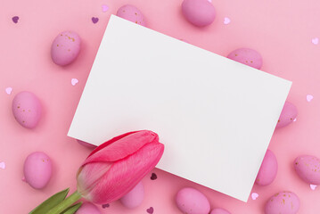 Easter postcard. Invitation mockup or blank with White card for greeting or invitation lies on Easter chocolate pink eggs. Red closed tulip. Shimmering glowing hearts. Happy holidays. Copy space