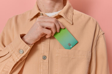 Anonymous man puts his green credit card in his pocket after payment on pink background copy space....