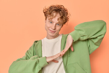 Portrait of a man with red curly hair and freckles in casual clothing green zipped hoodie and white shirt smiling and looking at camera while dancing and waving hands on orange backdrop copy space. 