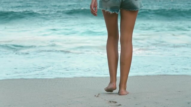 A woman's feet relax walking barefoot on the sand beach. Blue foam ocean water in background. Tanned slim girls legs in shorts. Travel, tourism, holiday. Beautiful nature summer landscape. Slow motion