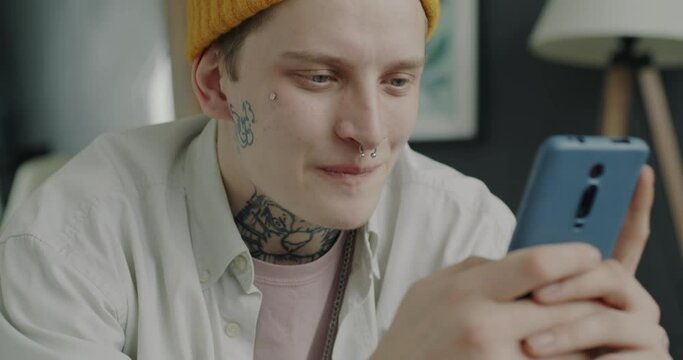 Young man with piercing and tattoo using smartphone touching screen and smiling at home. Online communication and generation Z people concept.