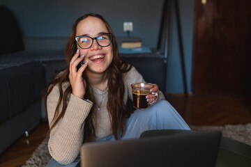 A young girl sitting at home while using laptop to study or do business while drinking coffee and using mobile phone