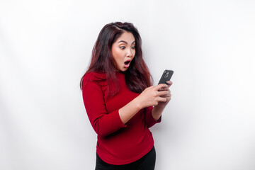 Fototapeta premium Shocked Asian woman wearing red top, holding her phone, isolated by white background