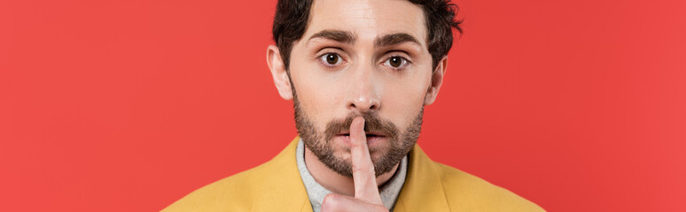 Obraz premium man in yellow blazer holding finger near lips while showing hush sign isolated on red coral background, banner.
