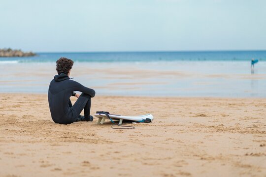 Curly-haired surfer sitting on the beach sand and looking at the seascape captured from behind