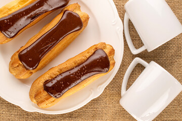 Three chocolate eclairs with a white ceramic plate and two cups on a jute cloth, close-up, top view.