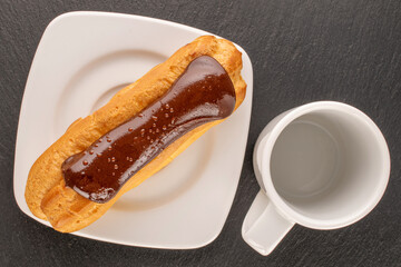 One chocolate eclair with white ceramic saucer and cup, close-up, on slate stone, top view.