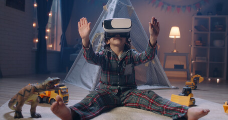 Funny little kid wearing pajamas is putting on virtual reality headset to dive into video games...