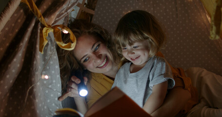 Happy caucasian mom and her cute baby boy spending time together at home, reading a book, using a flashlight in a cozy tent - happy family  © andreybiling