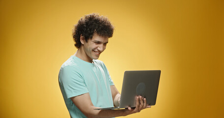 Close up shot of south asian guy with curly hair wearing casual clothing using his laptop, cheering about something and happily smiling, isolated on yellow background 