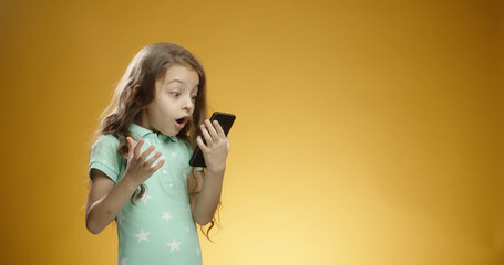 Funny Caucasian child holding and looking at phone, expressing an emotion of joy, surprised and shocked with open mouth isolated on a yellow background close-up. Copy space.