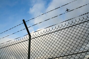 Plane flying against a blue sky with clouds over a chain link fence.