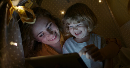 Positive caucasian woman and her son are watching cartoons on a tablet together, happily smiling. Family having fun in cozy tent in evening 