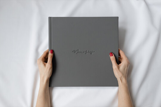 Close-up of a gray leather photo book with memories embossed in the hands of a girl with a red manicure on a white fabric background