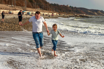 Father and daughter on the beach, running away from the incoming waves. The family jumps into the sea. Father and daughter are running on the waves of the surf. The concept of a friendly family. 