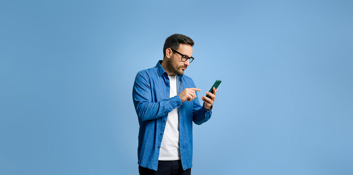 Serious handsome young adult man dressed in denim shirt surfing internet over smart phone while standing isolated against blue background