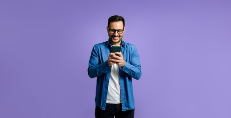 Charming smiling young adult man wearing blue denim shirt using social media and mobile phone while standing isolated on purple background