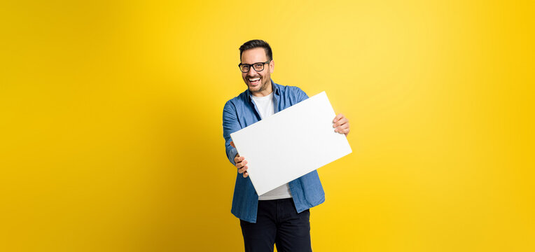 Cheerful male professional showing white blank banner for business advertising and looking at camera isolated on yellow background