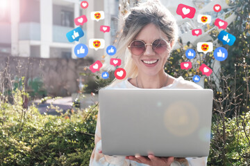 A happy blonde with a laptop gets emoji and emoticons for her online post. The concept of social...