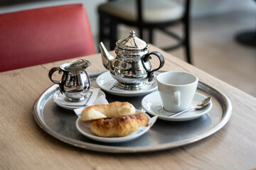 cafetera tetera de metal brillante cromada con taza pocillo de porcelana con platos redondos plancos sobre nadeja de metal, sobre mesa de madera en bar restaurante. Mediaslunas desayuno merienda