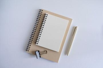 Top view of a blank page spiral bind notebook, A6 size on a white background on top of a kraft cover notebook, with a beige pen and black clip. Mock up notebook page.