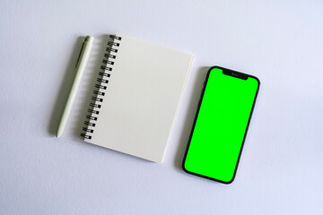 Apple smartphone mock up with green screen beside a notebook and a pen on a white background desk
