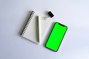 Apple smartphone mock up with green screen beside a notebook, a pen and a black clip on a white background desk