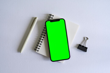Apple smartphone mock up with green on top of a notebook with a pen and a black clip on a white background desk