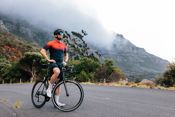 Fototapeta premium Young male cyclist relaxing after training with hands on a hips. Professional athlete with road bike enjoying the view while standing on a road in wild terrain.
