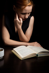 A woman is reading a book in the dark. Sits by the table.