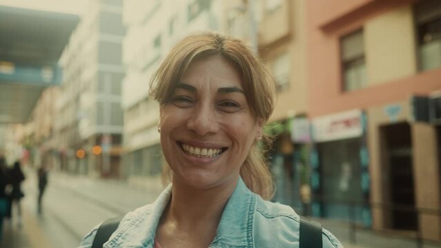 Happy Latin woman smiling in front of camera while waiting at tram station in the city