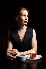 Woman at the table with tea and cake in the dark. Attractive forty-year-old woman in a black vest. Tea drinking. Loneliness.