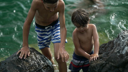 Small boy standing by rocks shore at lake water. Brave wet child stands outdoors holding on large stone
