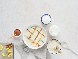 dessert of rice pudding with cinnamon powder on top along with the ingredients for the preparation: milk, sugar, cinnamon, lemon and rice. Top view on a pink marbled kitchen countertop.