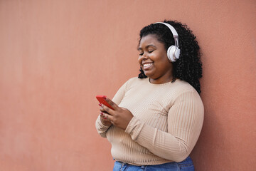 Happy curvy african girl using mobile phone while listening music playlist with wireless headphones