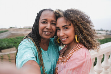 African mother and adult daughter taking a selfie while wearing traditional dress with ocean view...