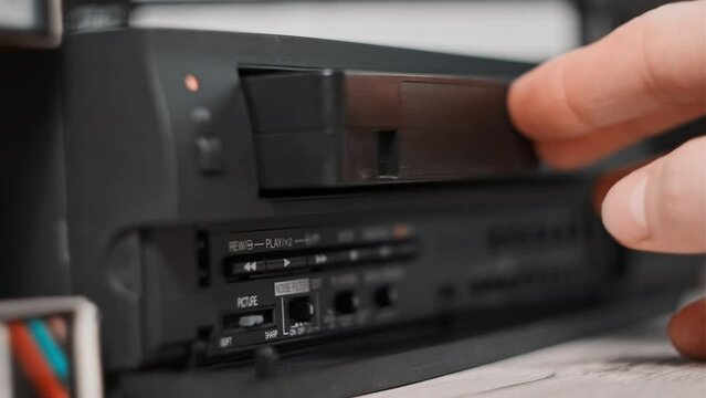 Male hand inserts VHS cassette into a VCR Video recorder. Black vintage videotape cassette recorder on desk with many video cassettes. Inserting retro VHS tape into vintage player. Home video archive