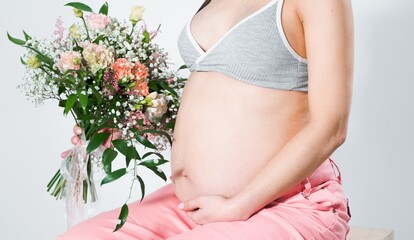 Pregnant woman with bouquet of flowers. Girl in gray top, pink pants holds hands on naked belly. Photoshoot of happy pregnancy, maternity, preparation. Baby expectation. Beautiful tender young mother