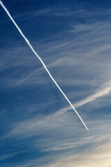 An airplane flying through a clear blue sky, leaving a long and persistent white contrail behind it. In the background, some soft and diffuse clouds add a touch of texture to the image. The bright blu