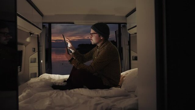 Young creative digital nomad working from his camper van at the seashore. He is using his tablet to remotely connect with colleagues. Beautiful sunset in the background. 