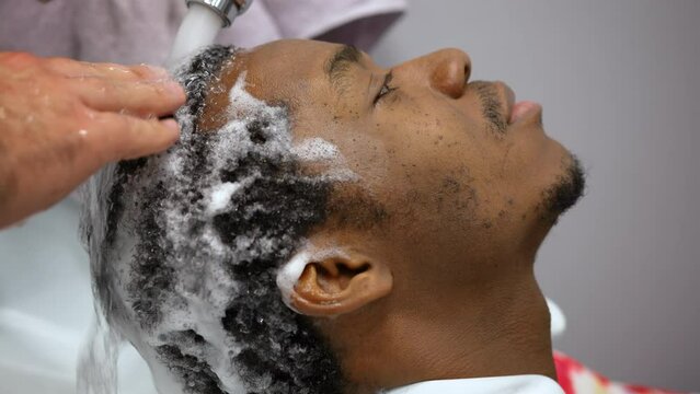 Barber is washing afroamerican man hair in barbershop pouring water, pside view.