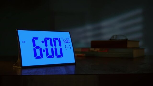 Digital alarm clock waking up at 6 AM. Close-up view. The numbers on the clock screen changes from 5:59 no 6:00 AM. Then alarm logo appears on the screen.
