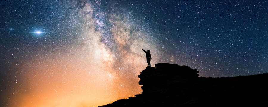 Lone person standing on top of rock, pointing with hand towards the Milky Way and the vast expanse of universe. Stunning silhouette of man gazing out into the cosmos. Power of dreams and aspirations.